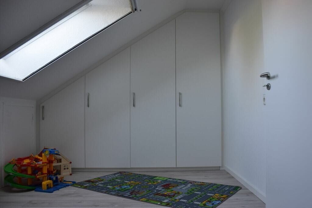 Une pièce avec un plafond en pente, des murs blancs, une porte dotée d’une poignée et un tapis de jeu coloré sur le sol.