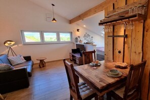 Ett mysigt vardagsrum med ett matbord i trä, stolar och en soffa. Ett fönster släpper in naturligt ljus och ger utsikt över grönområden utanför.