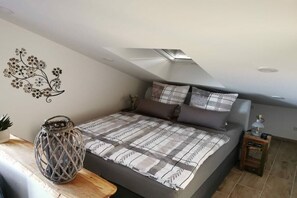 Een slaapkamer met een schuine plafond, een bed met een geborduurde deken, een wanddecoratie, een klein tafeltje met een plant en een nachtkastje met een lamp.