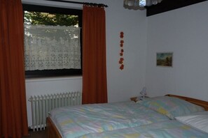 Ein Schlafzimmer mit einem Bett, einem Fenster mit Vorhängen, einem Heizkörper und einem kleinen Nachttisch mit Lampe.