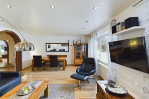Un salon moderne doté d’un téléviseur à écran plat, d’un fauteuil en cuir noir, d’une table basse en bois et d’un coin repas avec une table et des chaises en bois.