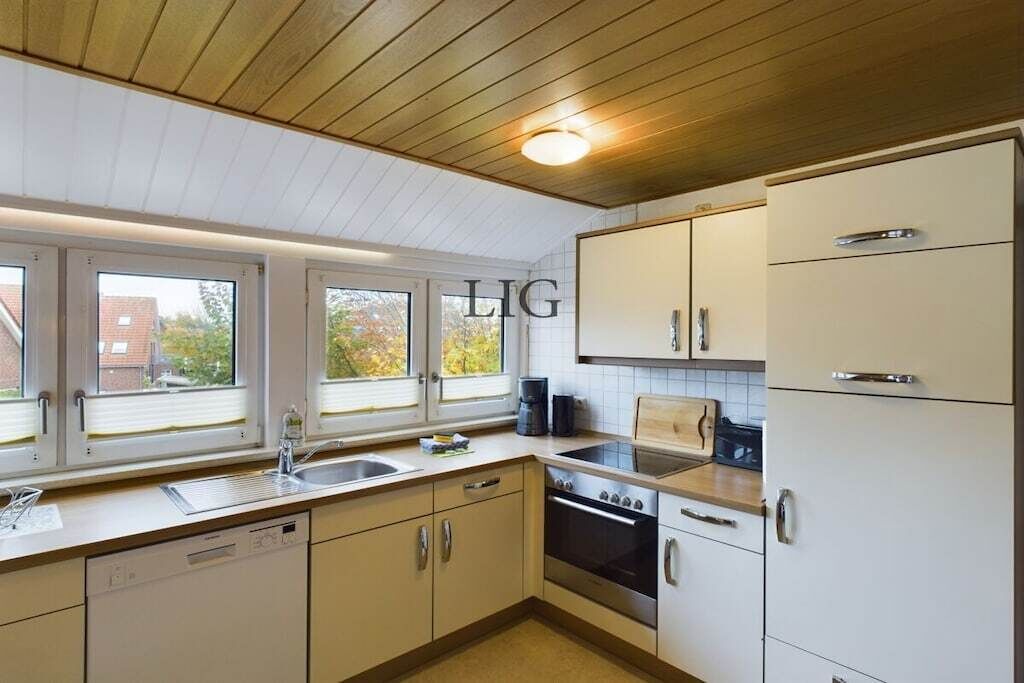 Een keuken met een vaatwasser, fornuis, oven en koelkast. Er zijn ramen met zonwering, een spoelbak en een aanrecht met een koffiezetapparaat.
