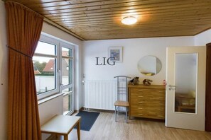 A room with a wooden ceiling, a window with curtains, a radiator, a desk, a chair, a dresser, and a mirror. - Holiday apartment Palladium 2 (Langeoog)
