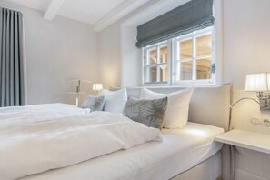 Una cama bien hecha con ropa blanca y almohadas decorativas, una mesita de noche con lámpara y una ventana con vistas al interior.