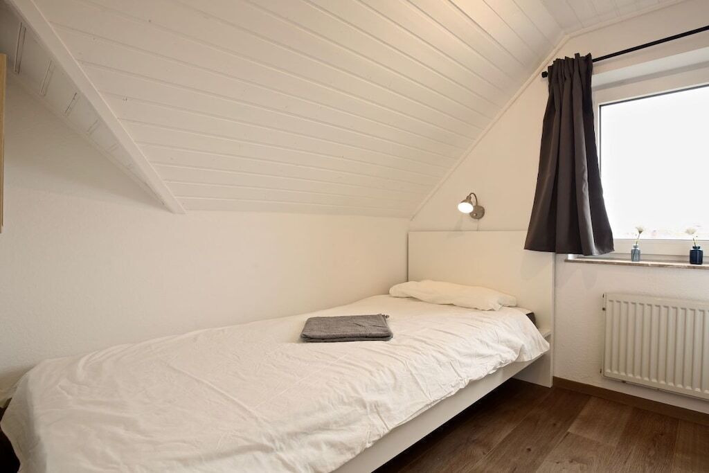 Een eenpersoonsbed met een witte matras en een grijze deken, een bedlampje, een raam met gordijn en een witte radiator.