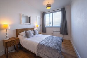 A bedroom with a bed, two bedside lamps, a mirror, a window with curtains, and a radiator. - Cosy former converted bank in heart of a busy working fishing village. (Mallaig)
