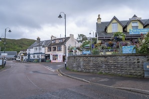 A street view with a row of buildings, including a cafe named 'The Tea Garden Cafe' and a sign for 'Backpackers Lodge'. - Cosy former converted bank in heart of a busy working fishing village. (Mallaig)