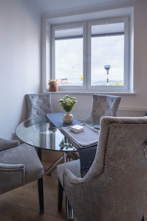 A dining area with a glass table, cushioned chairs, and a view through a window. - Cosy former converted bank in heart of a busy working fishing village. (Mallaig)