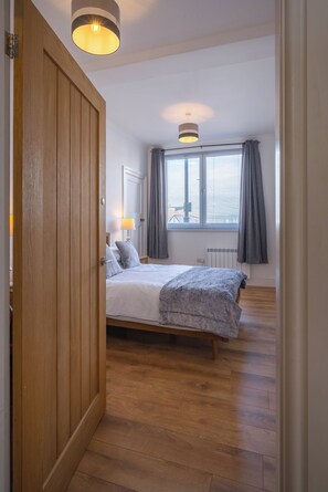 A bedroom with a wooden wardrobe, a bed with a grey blanket, a bedside table with a lamp, and a window with curtains.
