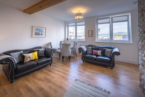 A living room with two leather sofas, a dining table, and chairs. There are large windows with a view of buildings and a cityscape. - Cosy former converted bank in heart of a busy working fishing village. (Mallaig)