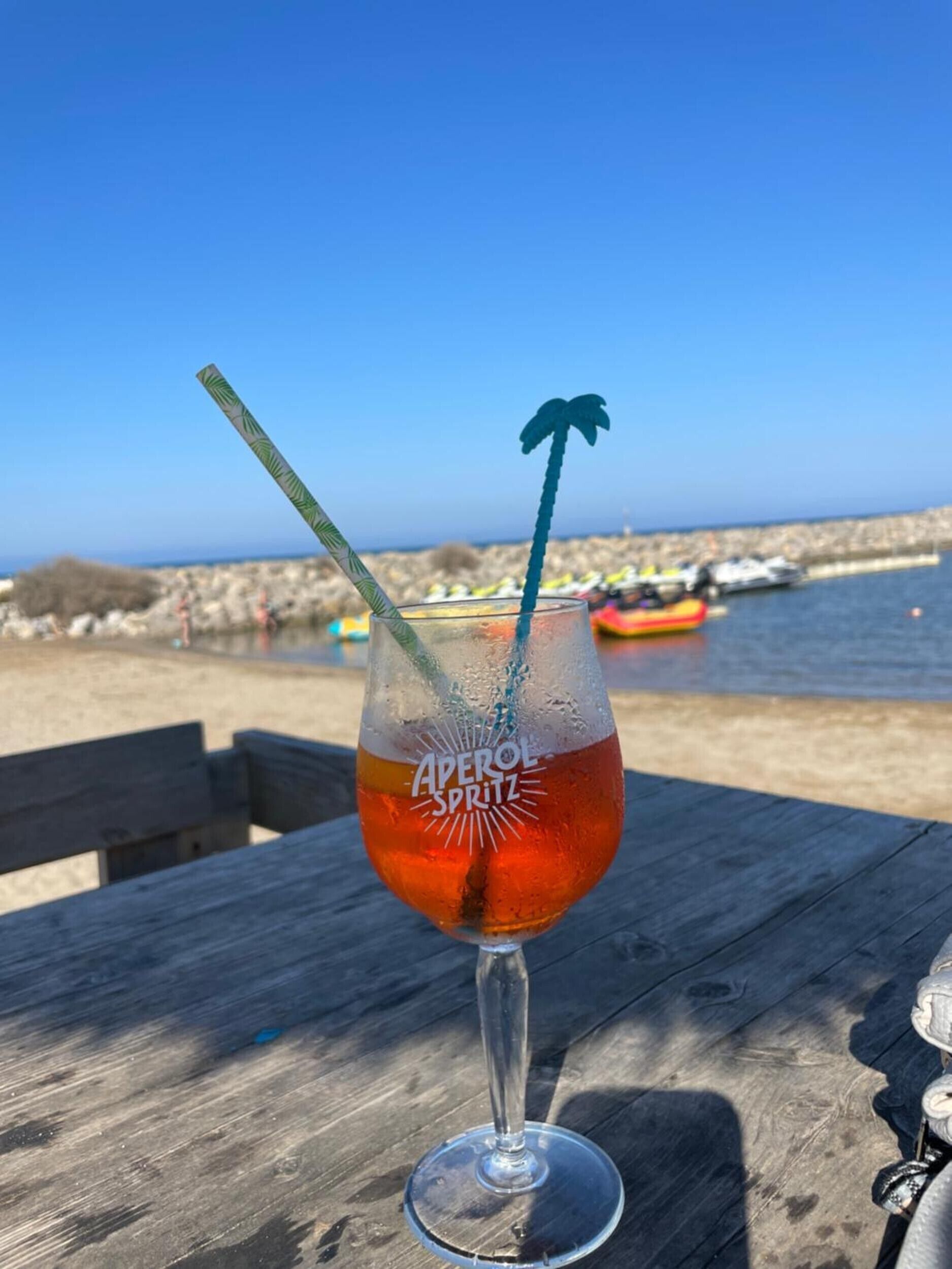 A glass of Aperol Spritz on a wooden table with a clear view of the sea and boats in the background.