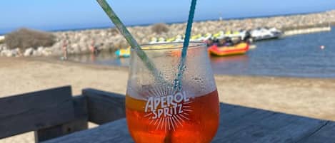 Un vaso de Aperol Spritz sobre una mesa de madera, con vista despejada al mar y barcos en el fondo.