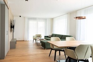 Ein modernes Wohnzimmer mit Holzboden, einem grünen Sofa, einem Esstisch mit Stühlen und einem an der Wand befestigten Fernseher.