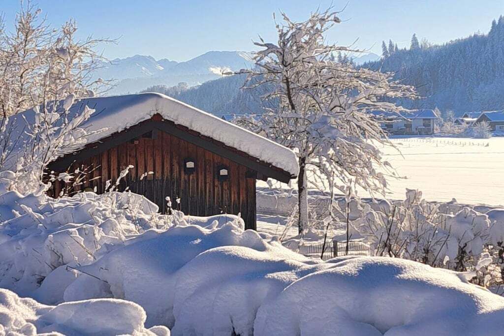 Eine Holzhütte mit schneebedecktem Dach, umgeben von schneebedeckten Bäumen und einer Bergkulisse.