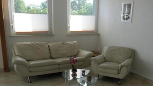 Un salon avec un canapé beige, une table basse en verre et un petit fauteuil. Il y a deux fenêtres avec des stores, un tableau encadré au mur et une plante en pot sur la table.