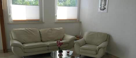 Un salon avec un canapé beige, une table basse en verre et un petit fauteuil. Il y a deux fenêtres avec des stores, un tableau encadré au mur et une plante en pot sur la table.