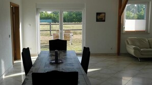 Un coin repas avec une table et des chaises, une porte-fenêtre coulissante donnant sur un jardin, et une fenêtre avec des stores.