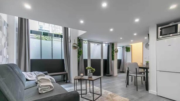 A modern living room with a sofa, a small table, and a kitchen area with a refrigerator and microwave. - Apartment Cosy - 2BR/6P - Near Montorgueil (Paris)