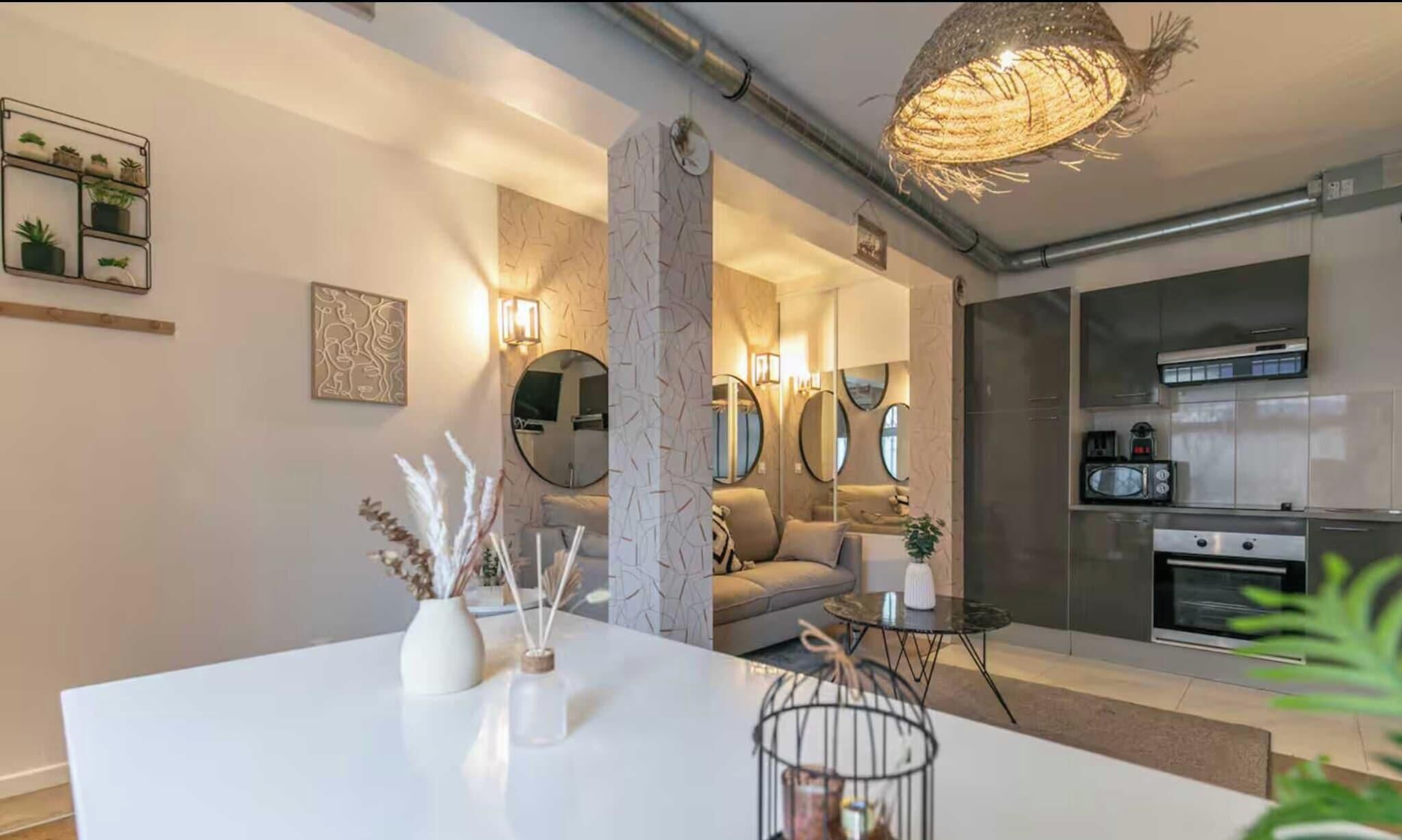 A modern kitchen with stainless steel appliances, a dining area with a white table, and a living space with a sofa and decorative mirrors.