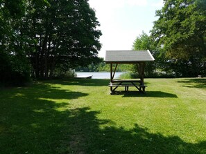 A picnic table with a roof, surrounded by grass and trees, with a body of water in the background.
