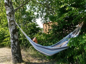 Un hamac est suspendu entre deux arbres, dans un environnement naturel caractérisé par un feuillage dense, avec un bâtiment visible au loin.