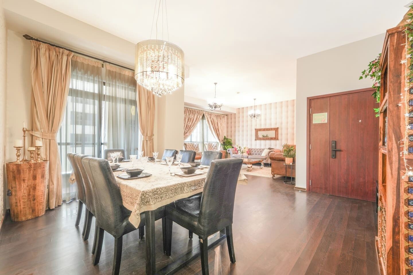 Een eetgedeelte met een ronde tafel, stoelen en een kroonluchter. Achtergrond toont een bank en een deur.