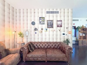 A living room with a brown leather sofa, a round side table with a lamp, and a wall decorated with various framed items and a clock. - Spacious 3BR in Amwaj | Steps from the Beach (Dubai)
