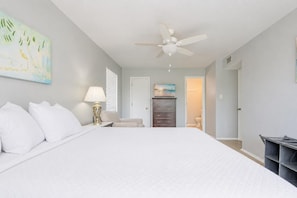 A bedroom with a large bed, a ceiling fan, a nightstand with a lamp, a dresser, and a painting on the wall. - Romar Village 312  Family-Friendly Beach Retreat (Orange Beach)