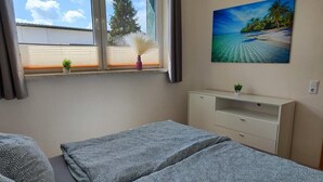 Quarto com duas camas, janela com persianas, pintura de uma praia e uma pequena planta no criado-mudo.