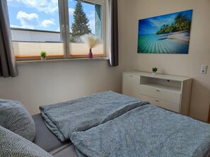 A room with two beds, a window with blinds, a painting of a beach, and a small plant on the dresser. - Cozy Holiday Apartment with Private Garden (Milmersdorf)