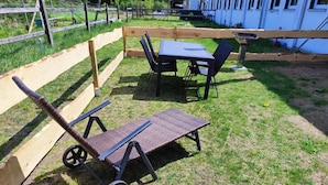 Uma área externa com gramado, cercada por um muro de madeira, e uma mesa com cadeiras, incluindo uma cadeira de rodas.