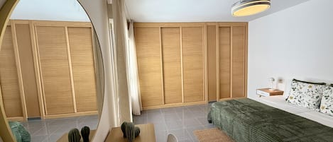 A modern bedroom with a wooden wardrobe, a round mirror, a green sofa, and a bedside table with a lamp.