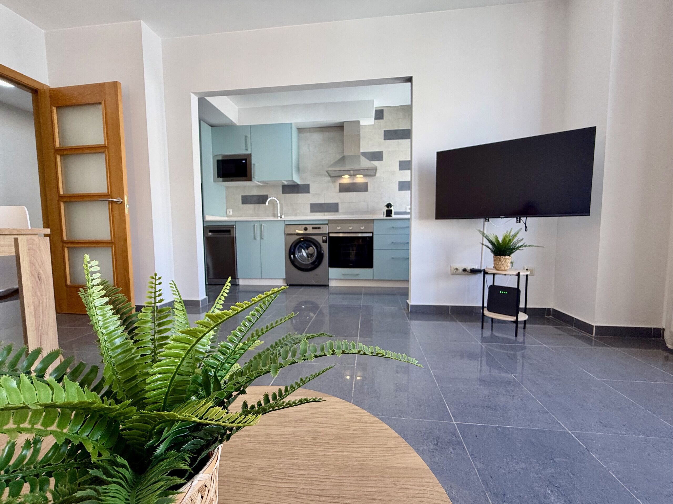 Una cocina moderna con lavadora, un televisor de pantalla plana montado en la pared y un helecho grande en primer plano.