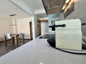 A modern kitchen with a built-in espresso machine, a dining area with a wooden table and chairs, and a contemporary light fixture. - Dragos Suites 201 (Arrecife)