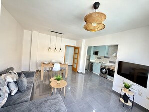 A modern living room with a sofa, a dining table with chairs, a kitchen area with appliances, and a TV mounted on the wall. - Dragos Suites 201 (Arrecife)