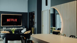 A modern living room with a large flat-screen TV displaying 'NETFLIX,' a black leather sofa, a dining table with chairs, and a decorative vase with flowers. - Top Provenza Penthouse 3-bedroom apartment w/ AC, Elevator, Fast Wifi, Parking (Medellín)