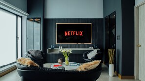 Ein modernes Wohnzimmer mit einem großen Flachbildfernseher, der 'NETFLIX' zeigt, einer schwarzen Ledersofa, einem kleinen runden Couchtisch mit Büchern und einer Blumenvase, sowie einer Glastür zum Garten.