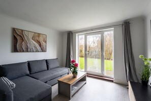 A modern living room with a grey sofa, a wooden coffee table, and a large window with curtains. - Karazo Ltd - 2 bedroom home in Croydon (Croydon)