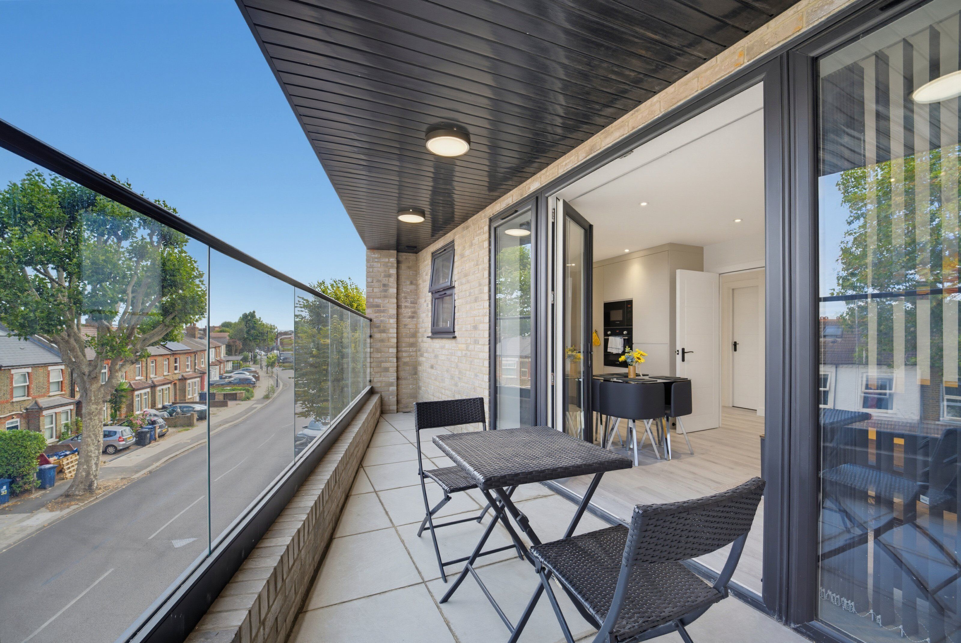 En balkon med et bord og stole, udsigt til gaden, og et kig ind til et køkkenområde.