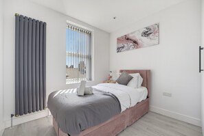 A modern bedroom with a bed, a radiator, a window with blinds, and a piece of wall art. - 2-Bed with Balcony in Northfields -Apt 4 (London)