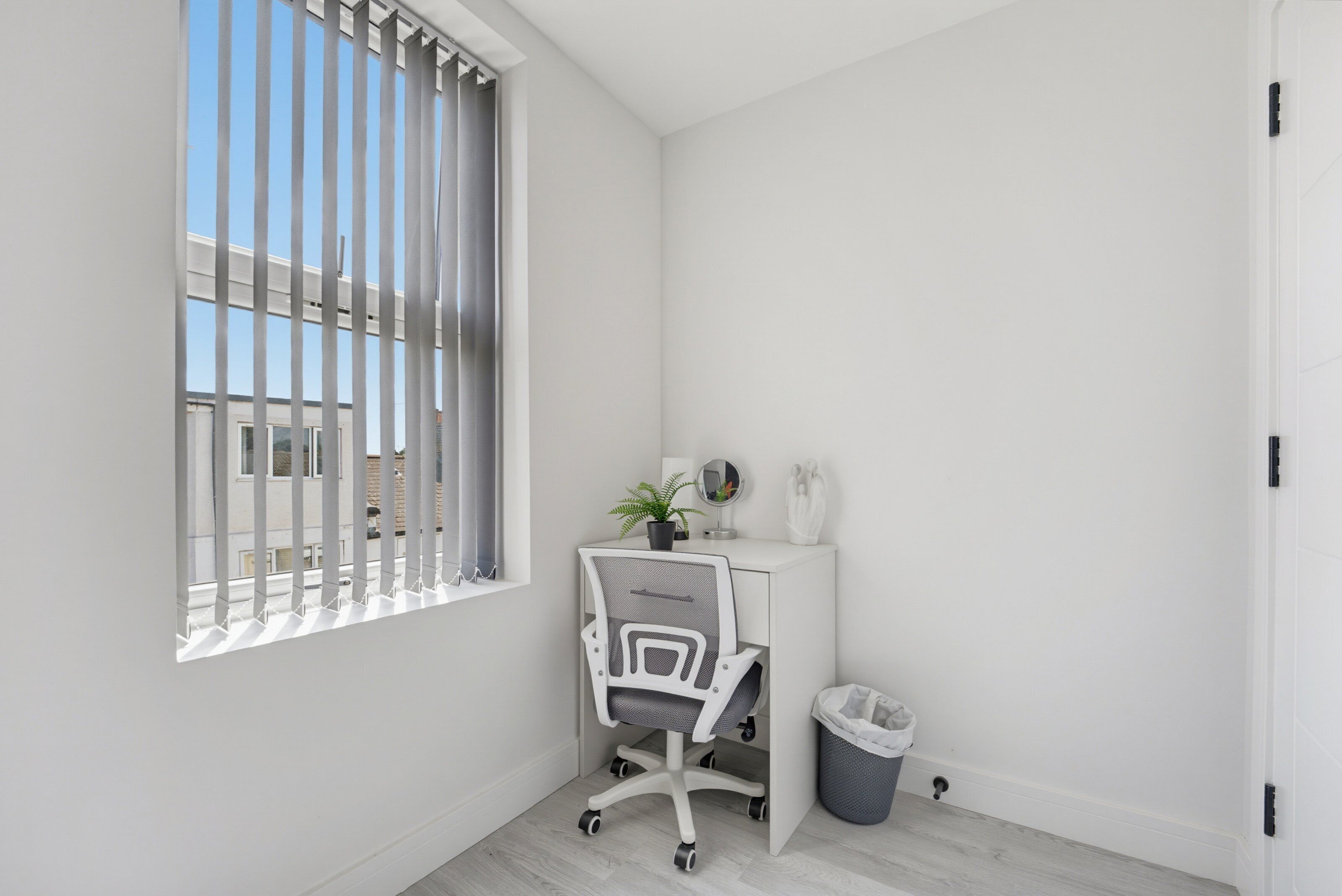 Quarto pequeno e branco com escrivaninha, cadeira e janela.