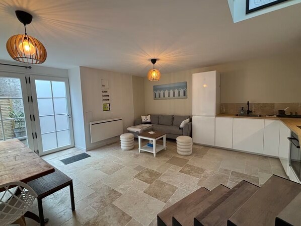 A modern kitchen with white cabinets, a dining area with a wooden table, and a sofa. - Vrbo Property (Saint-Vaast-la-Hougue)
