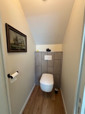 A small bathroom with a toilet, a framed picture on the wall, and a roll of toilet paper. - Vrbo Property (Saint-Vaast-la-Hougue)