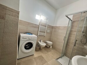 A compact bathroom with a washing machine, toilet, and glass-enclosed shower. - TRULLO PATANID, located in the heart of the Itria Valley, in a strategic location (Puglia)
