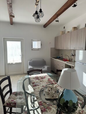 A dining area with a glass table, chairs, and a tray with teacups and saucers. There is a window with a view of trees, a door, and a kitchen area with cabinets and a refrigerator. - TRULLO PATANID, located in the heart of the Itria Valley, in a strategic location (Puglia)