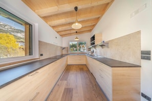 A modern kitchen with wooden beams, a long countertop, and a view of a mountain landscape.