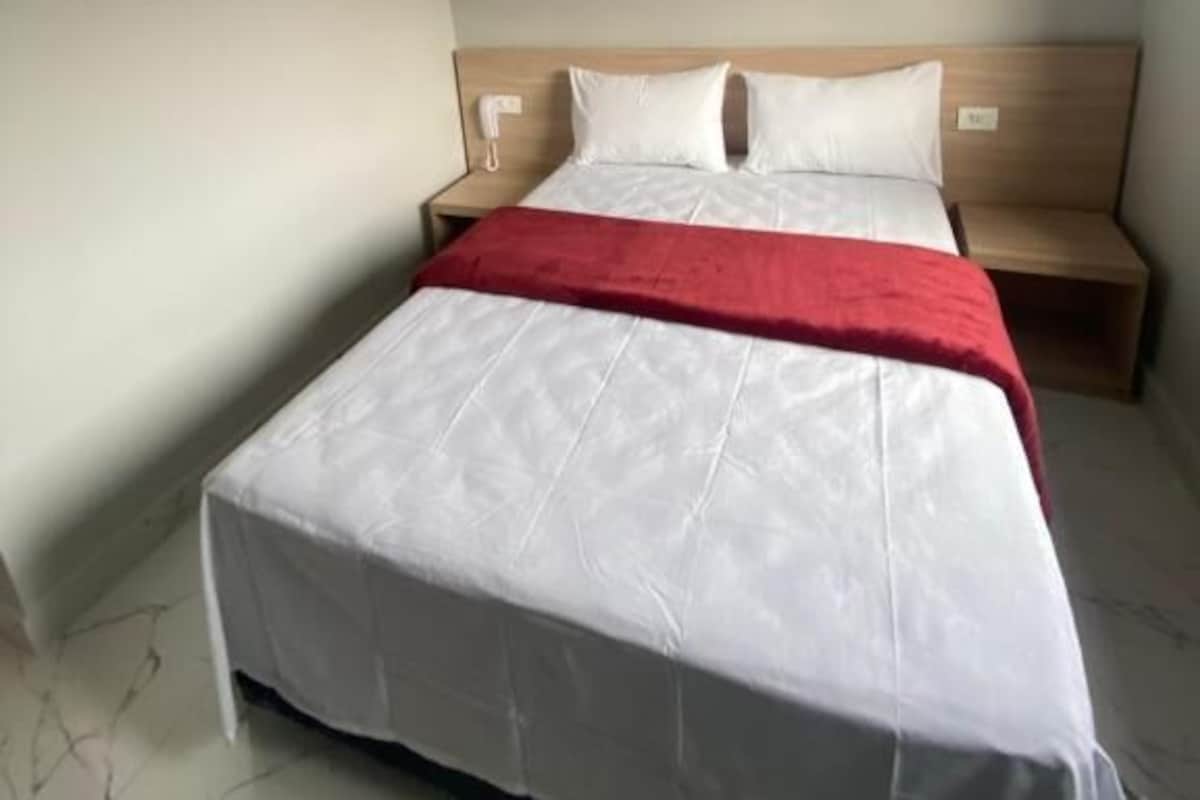 Una cama individual con cabecera de madera, una colcha roja y un edredón blanco en una habitación con suelo de mármol y un equipo de aire acondicionado instalado en la pared.