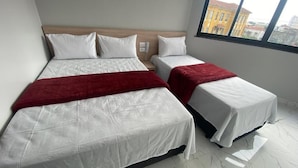 Habitación de hotel con dos camas, cabecera de madera, manta roja y vista a la ciudad a través de un ventanal grande.