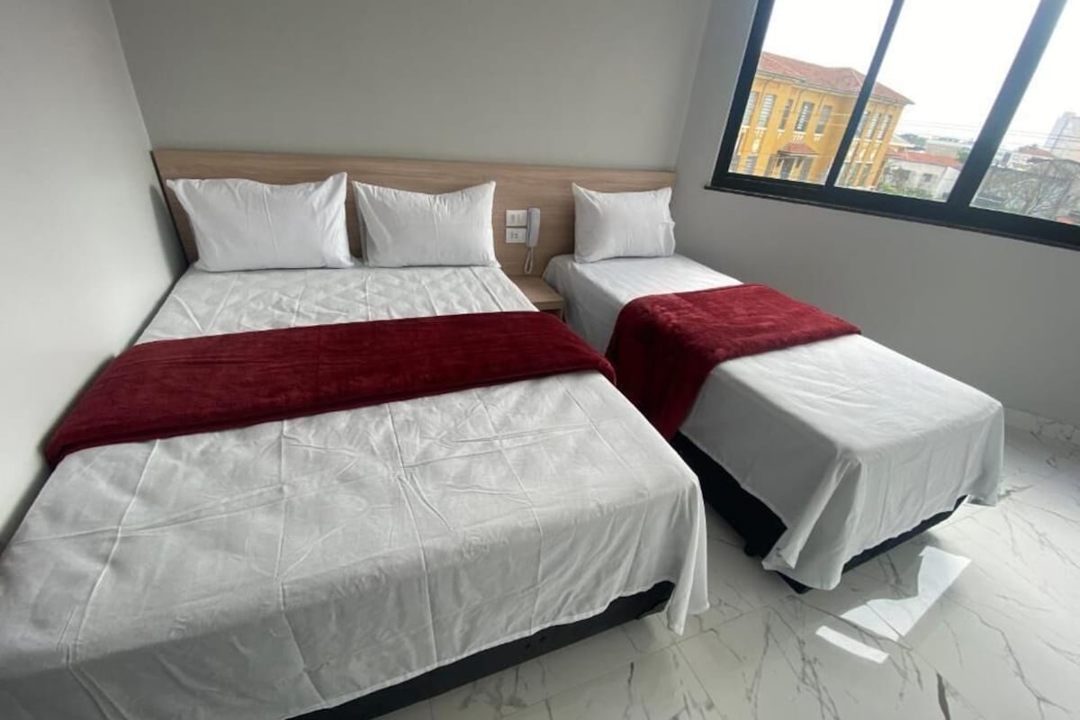 Habitación de hotel con dos camas, cabecera de madera, manta roja y vista a la ciudad a través de un ventanal grande.