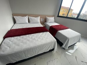 A hotel room with two beds, a wooden headboard, a red throw blanket, and a city view through a large window.